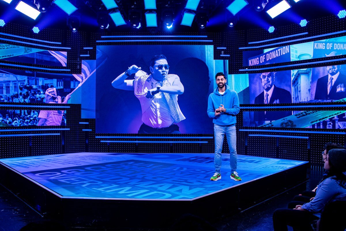 Hasan Minhaj mengenakan hoodie biru berdiri di pinggir panggung Patriot Act ketika foto Psy tampil di layar